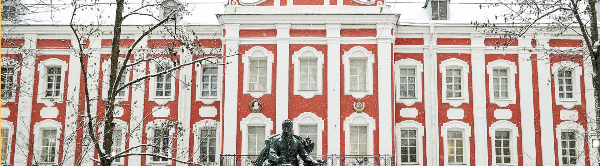 Foto a da fachada da Universidade de São Petersburgo na Rússia. Prédio vermelho com janelas, colunas e beirais brancos, uma estátua de um homem a frente do edifício e o chão da parte exterior coberto por neve.